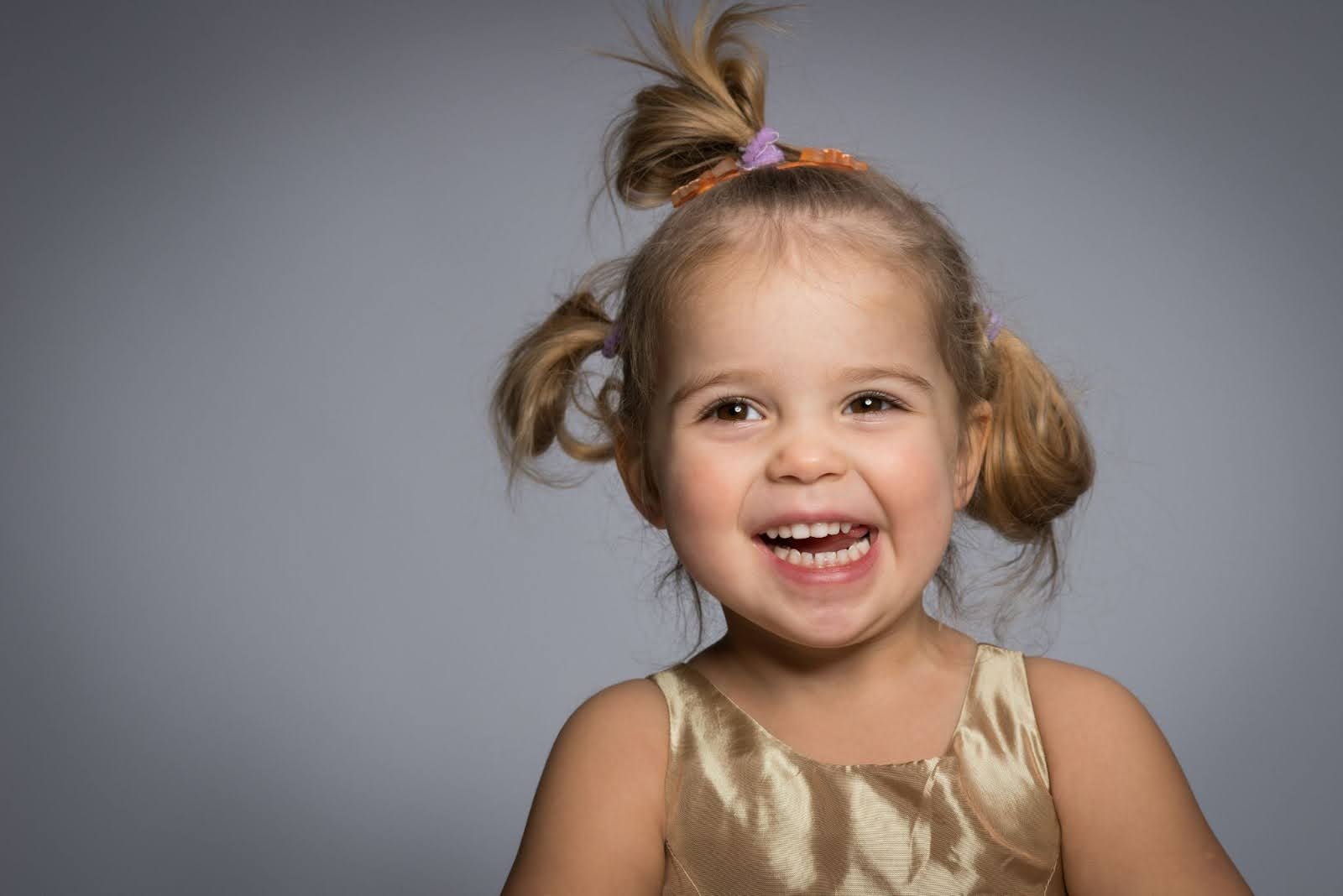 Toddler in a gold top with messy buns in her hair smiles against a gray background