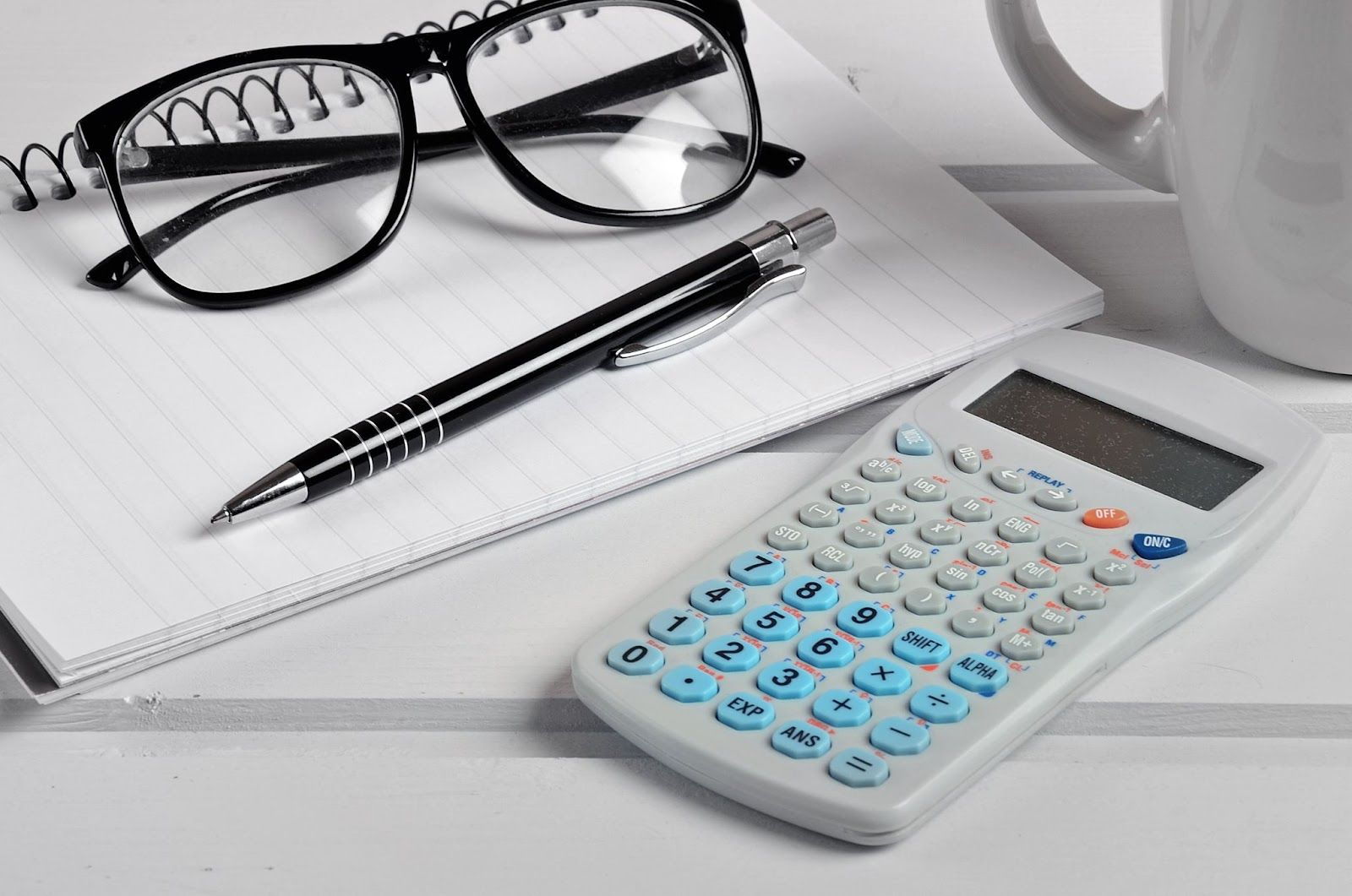 Glasses, pen, spiral notebook, calculator, and coffee mug rest atop a white surface