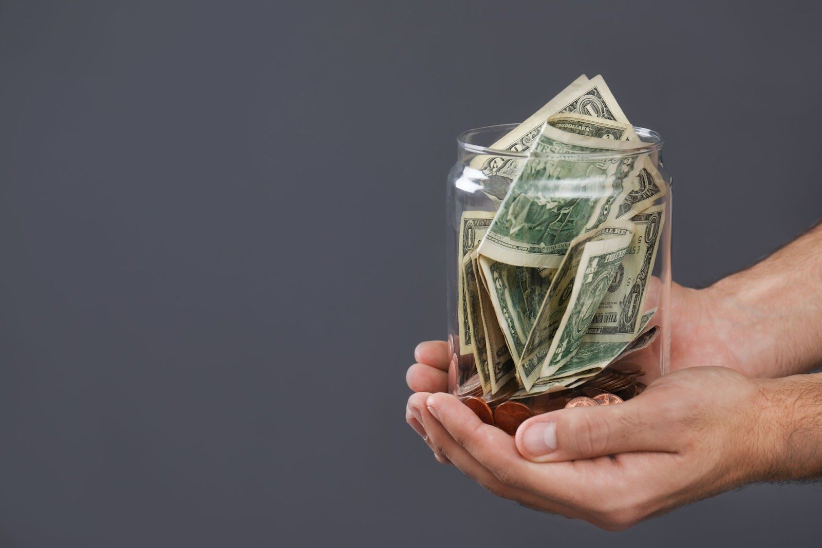 Cupped hands hold glass jar of dollar bills, against a gray background