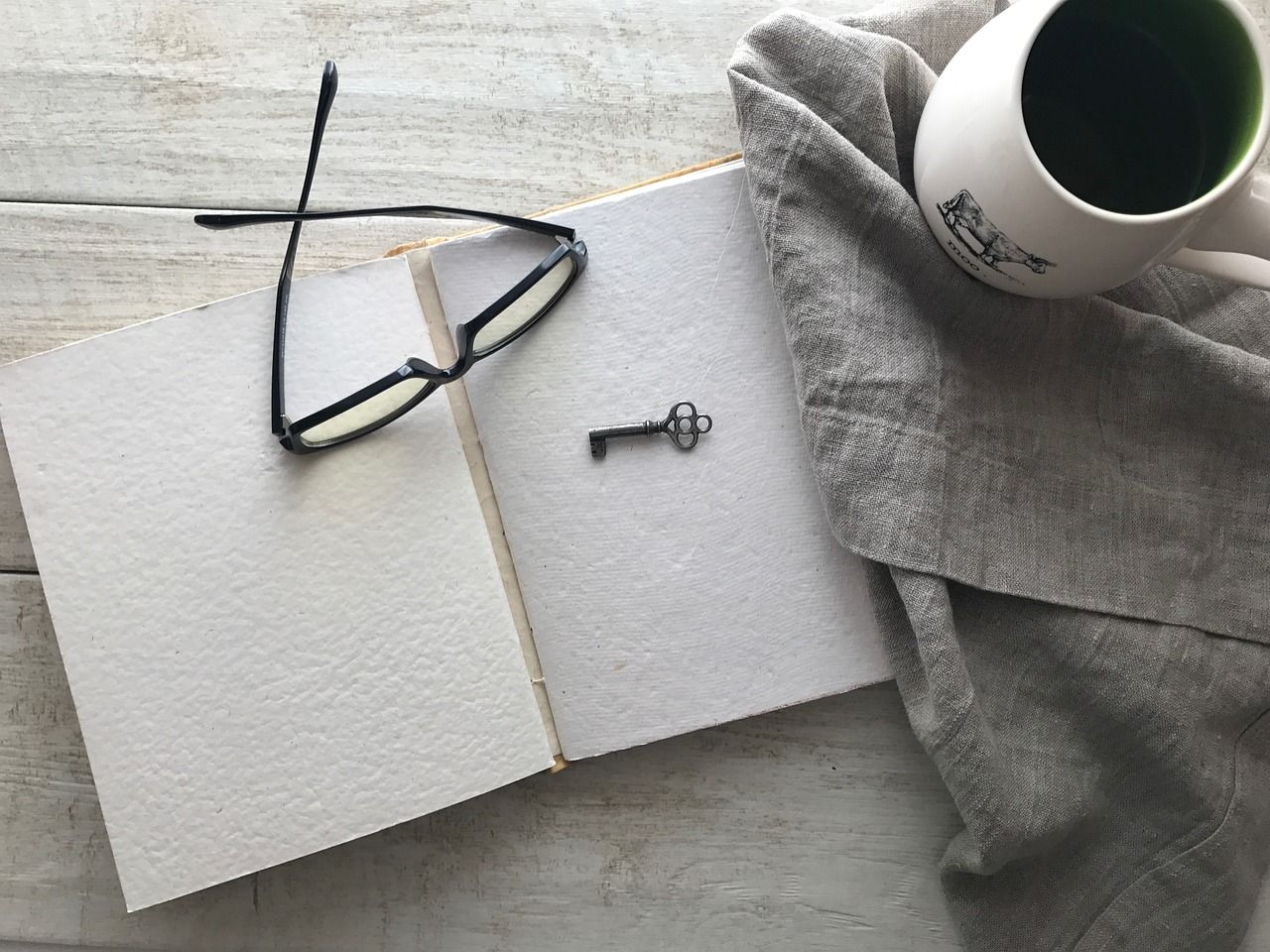 Open notebook with glasses and vintage key on top, beside a mug a of coffee
