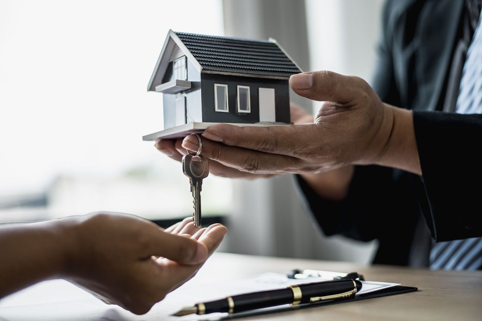 Hands holding a model gray house lower a house key into another person's open palm