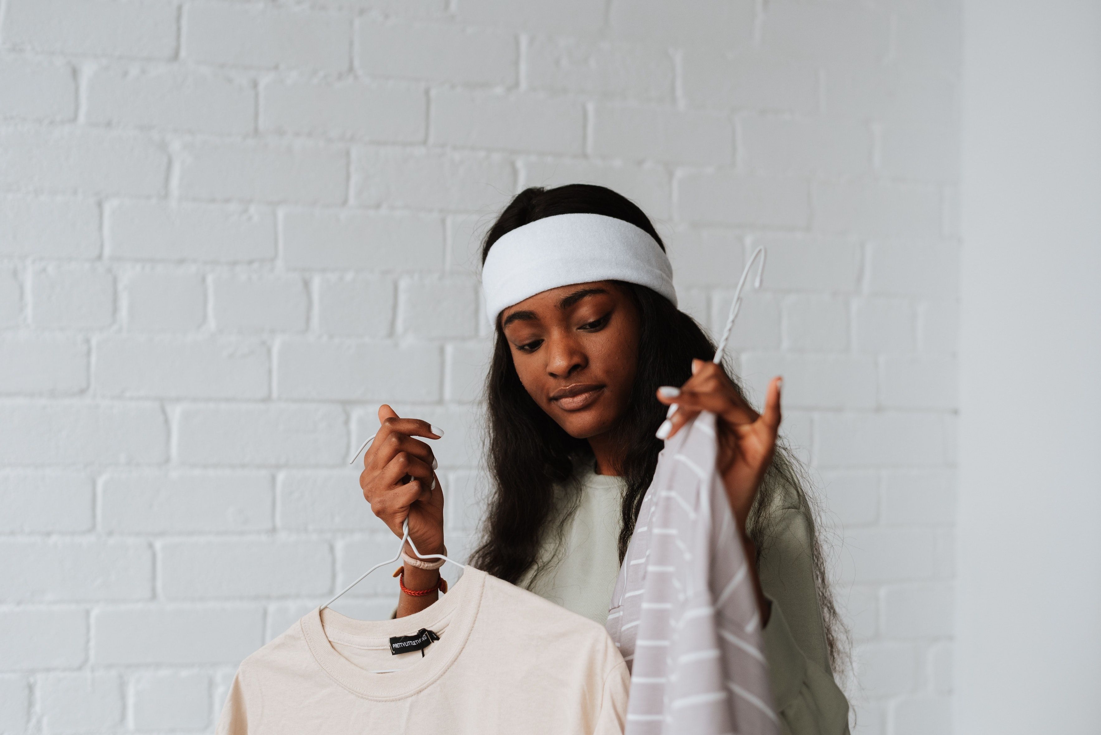 In a white-brick room, a girl in white headband holds up two pieces neutral-colored of clothing