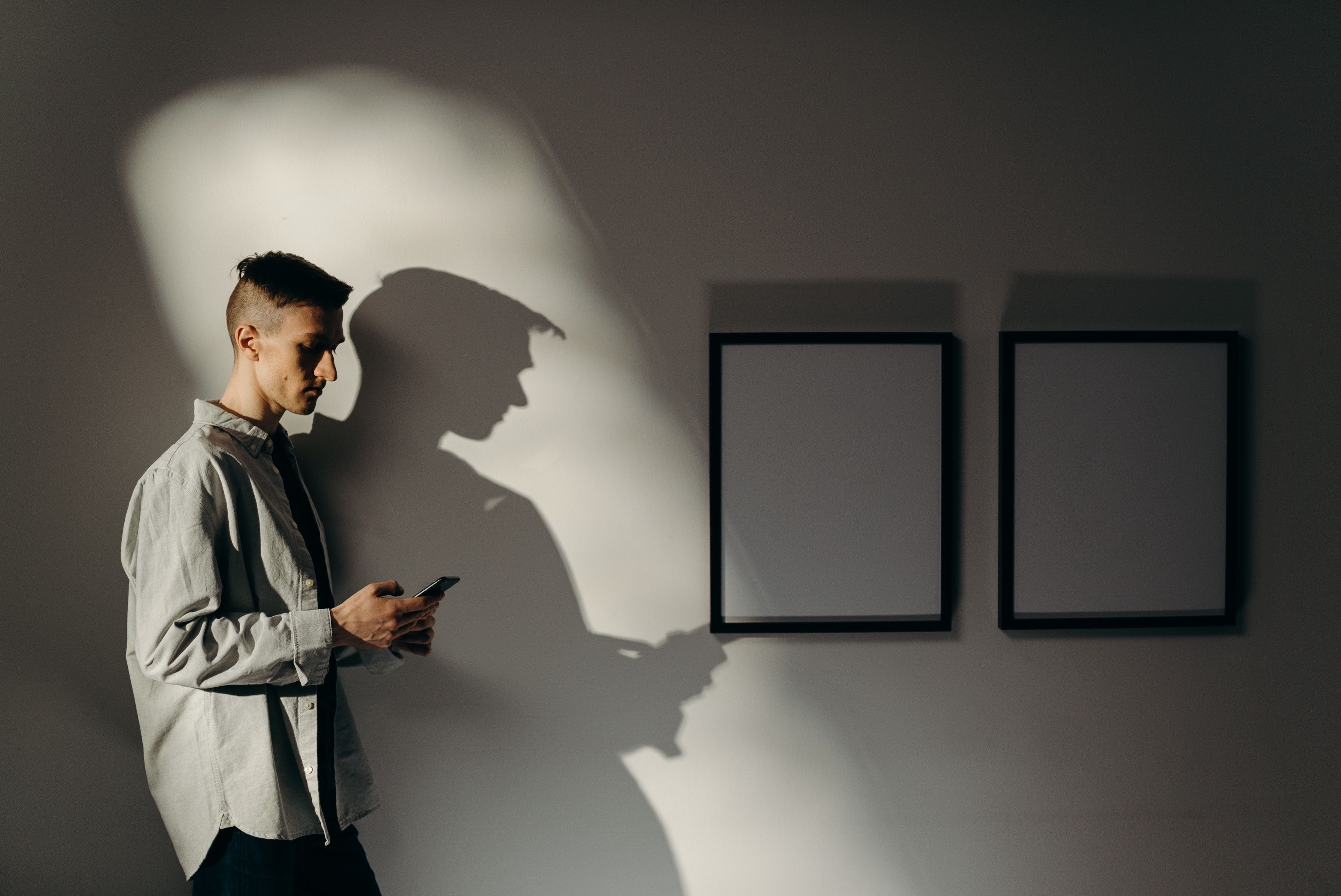 In a dark, window-lit room, a man in collared shirt looks down at his phone beside two empty wall frames