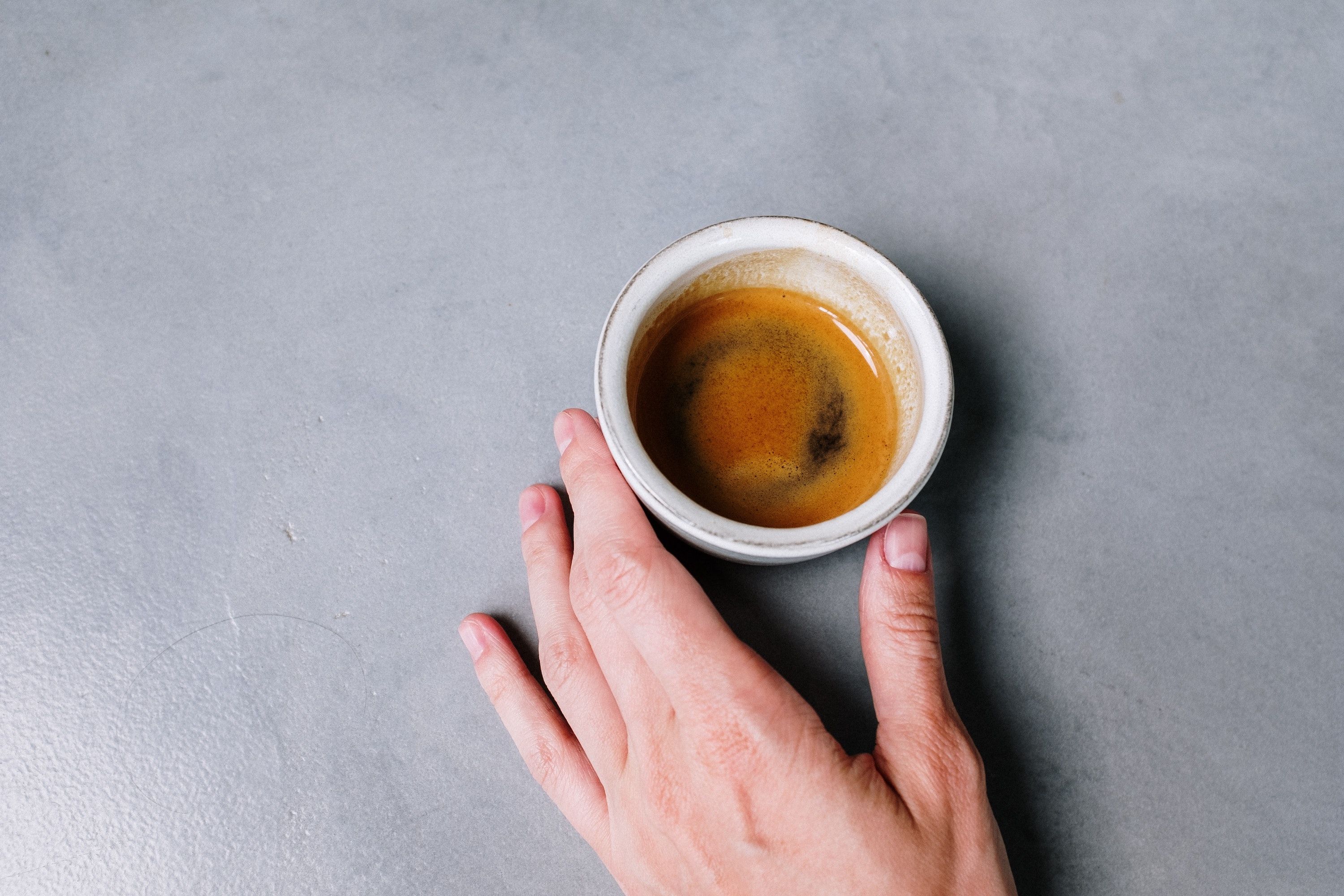 Hand holds lip of espresso cup on gray surface