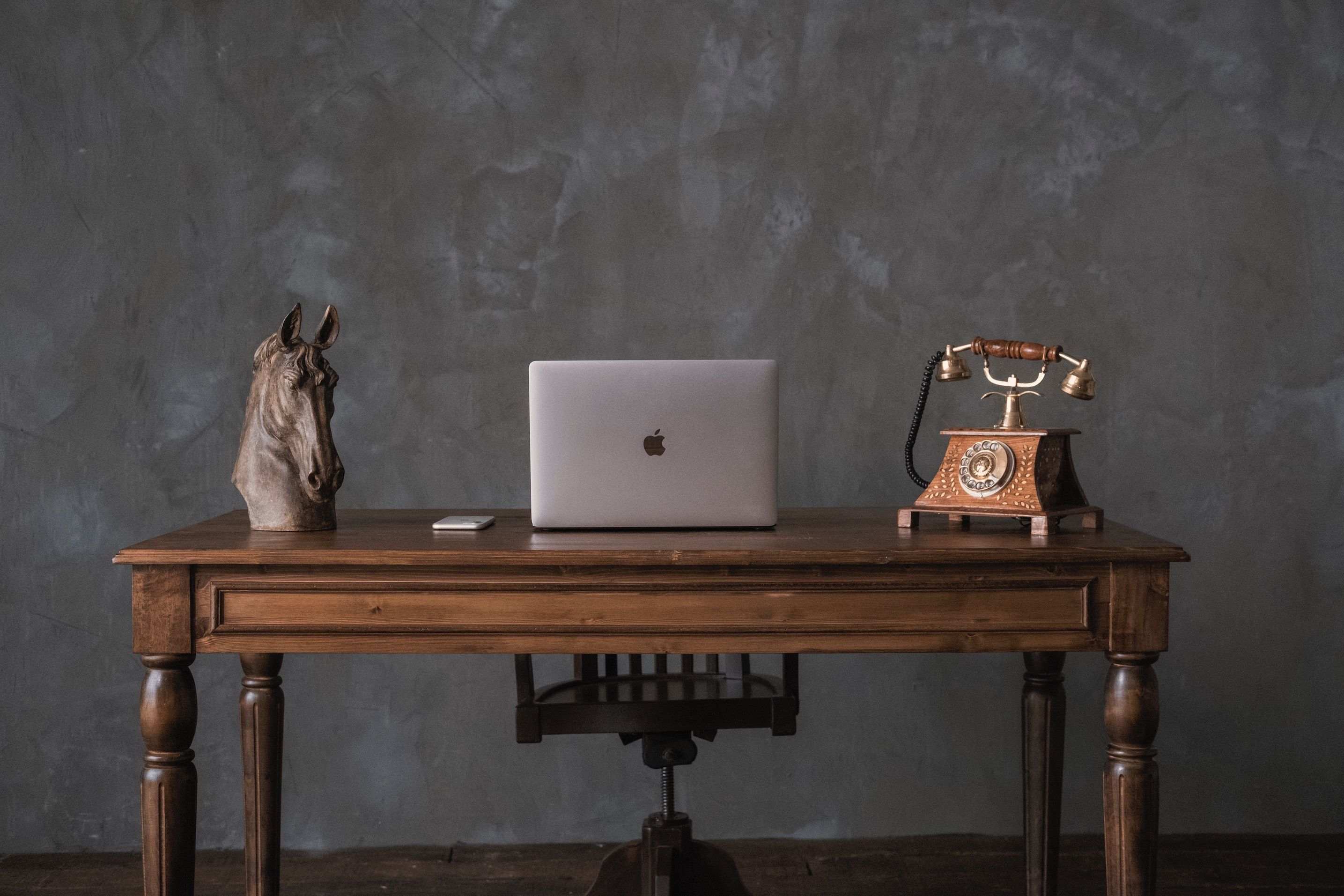 Horse-head sculpture, MacBook, and dial phone atop traditional wooden desk in a dark gray room