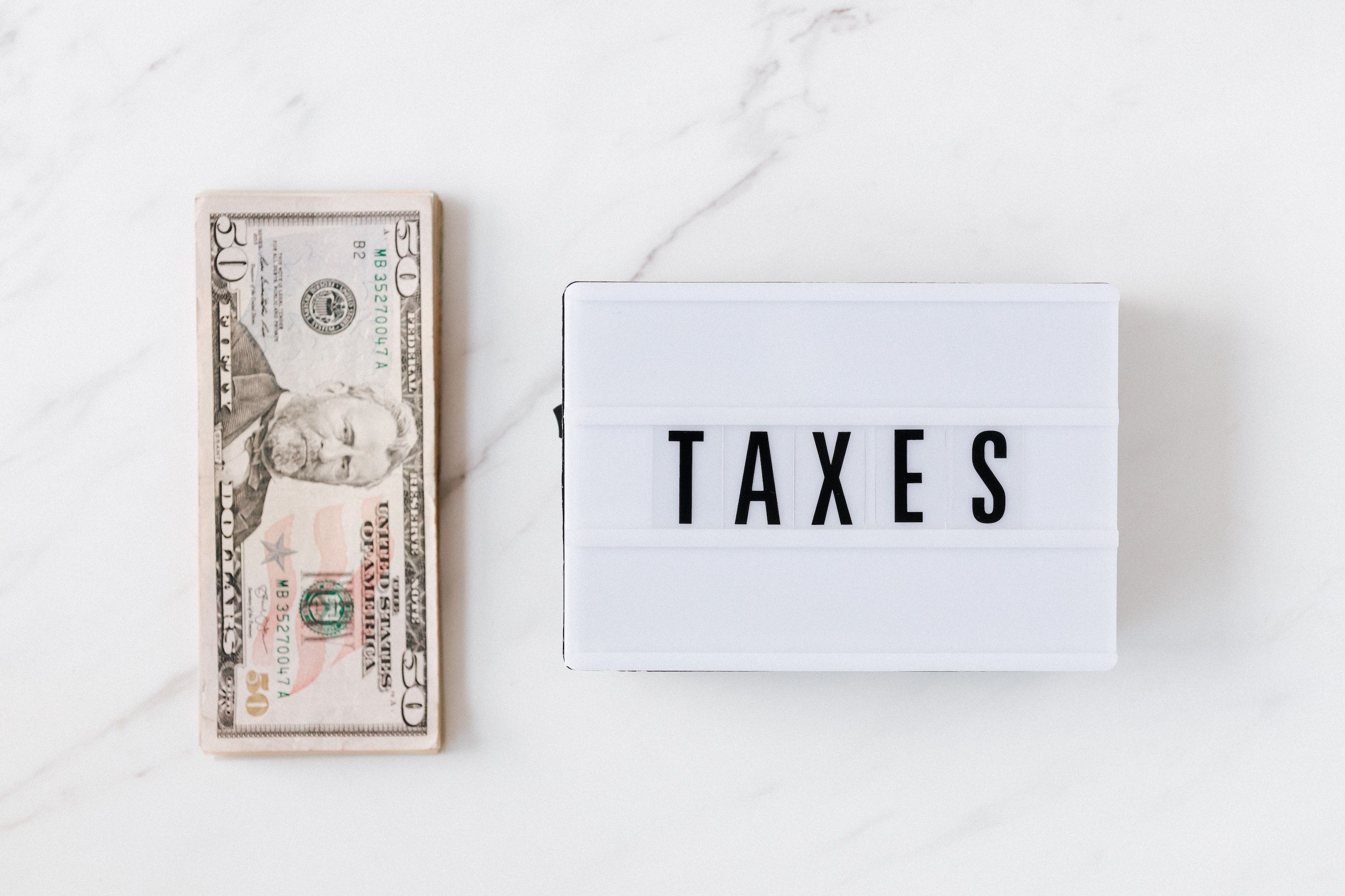 Flat lay of stack of bills and sign that says "TAXES," on a white marble surface