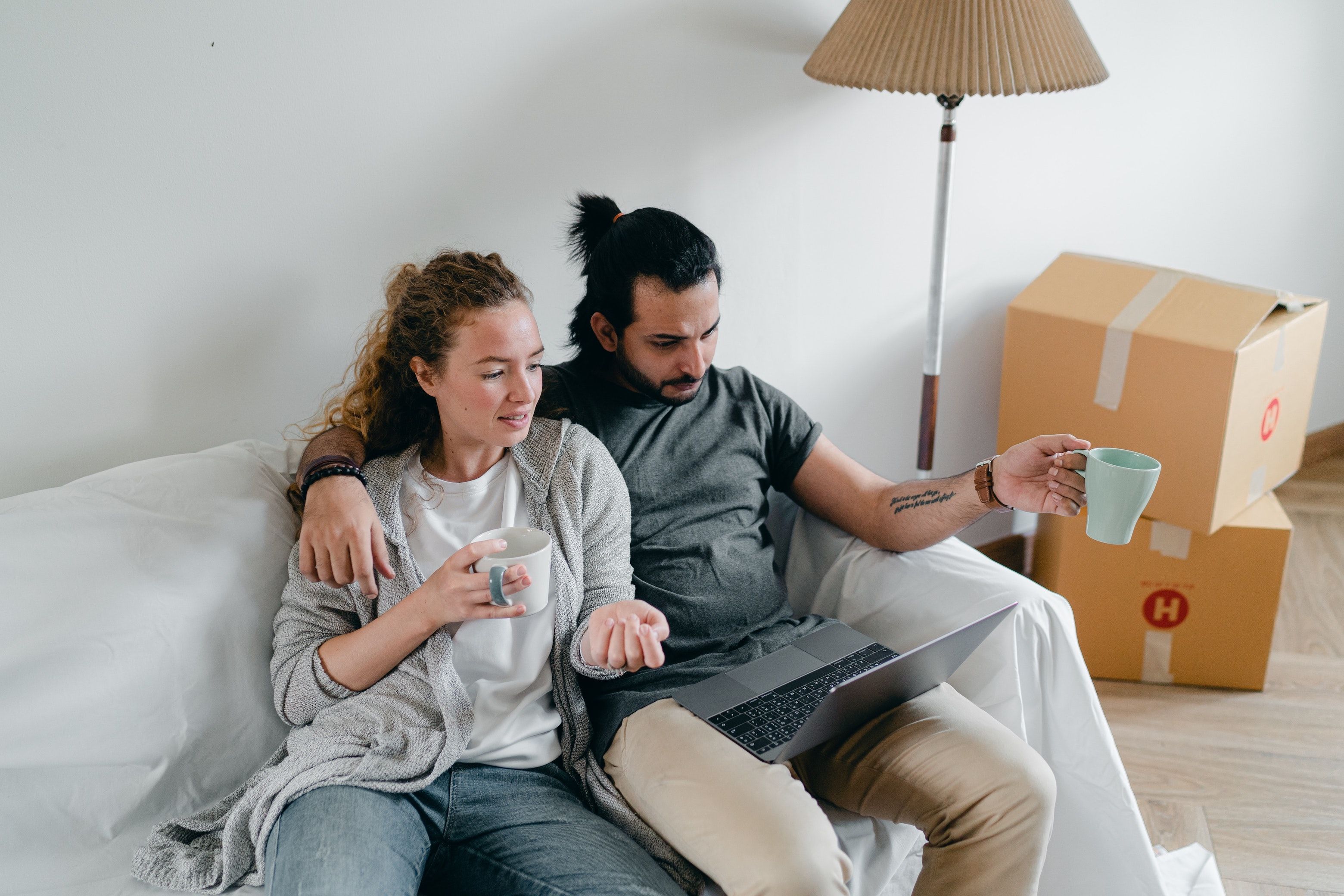 Couple sits in bare apartment with moving boxes, looking at a laptop and drinking coffee