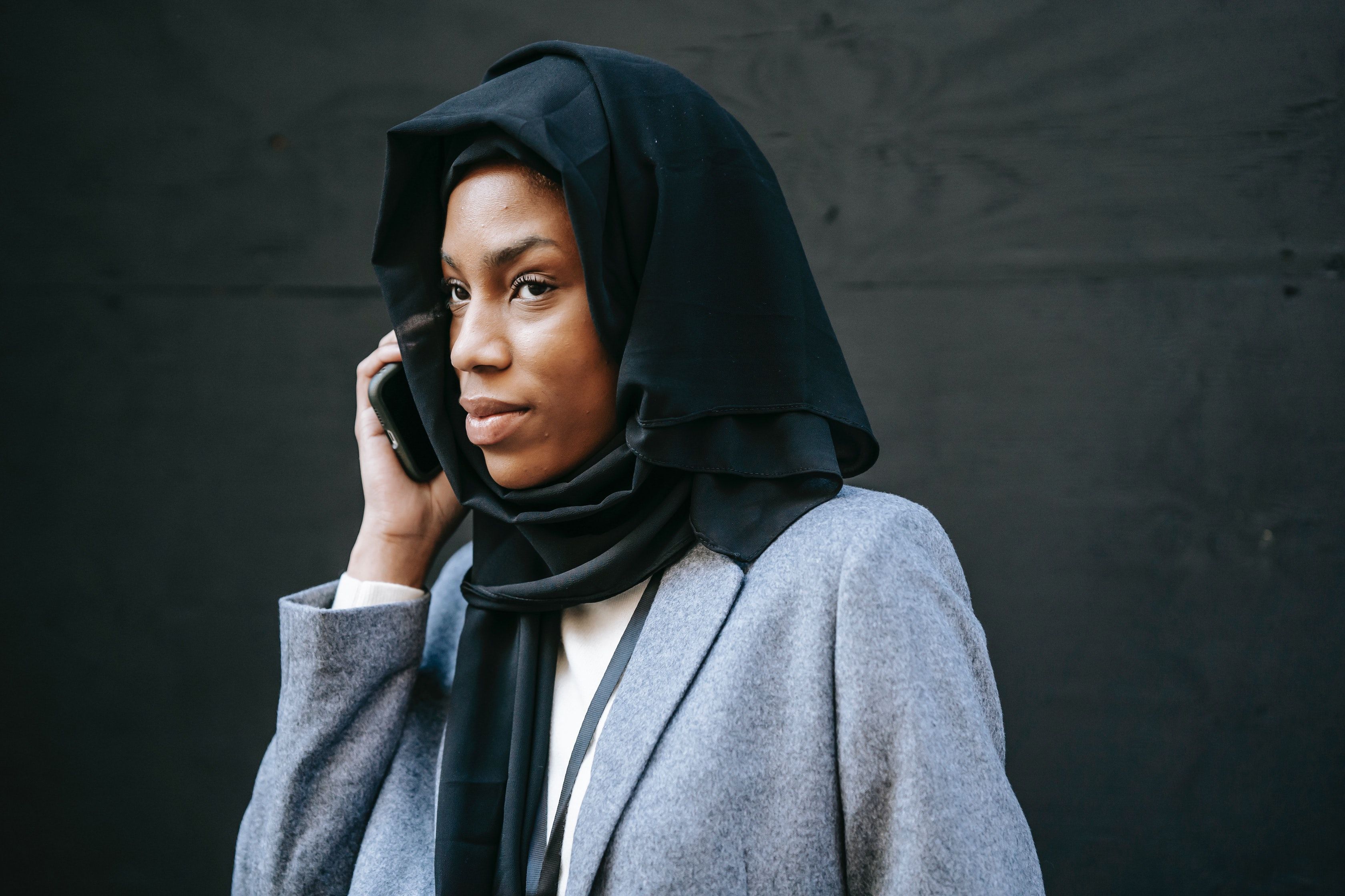 Young woman in blazer and headscarf holds cell phone to her ear, against a dark gray wall