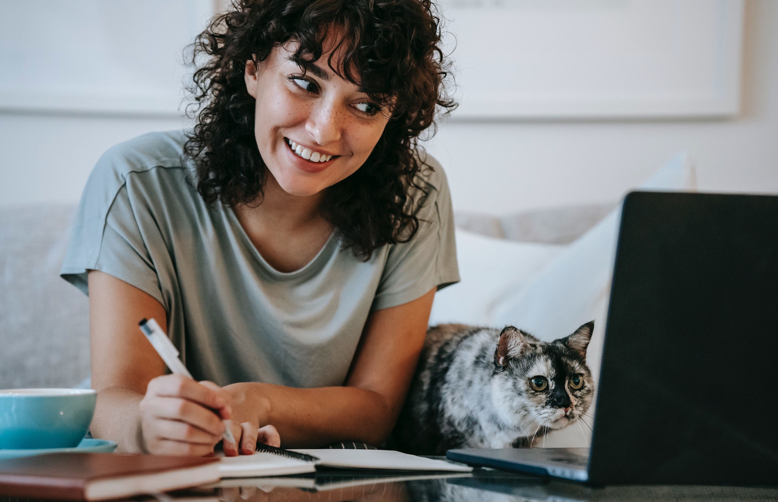 Smiling woman with curly hair in gray shirt writes in a notebook, laptop open on the table and cat by her side